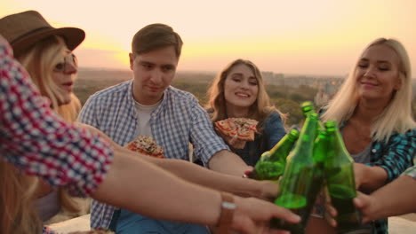 Young-men-and-women-clinks-glasses-and-drinks-beer-from-green-bottels-on-the-party-with-friends-on-the-roof-at-the-sunset.They-eat-hot-pizza-after-in-summer-everning.